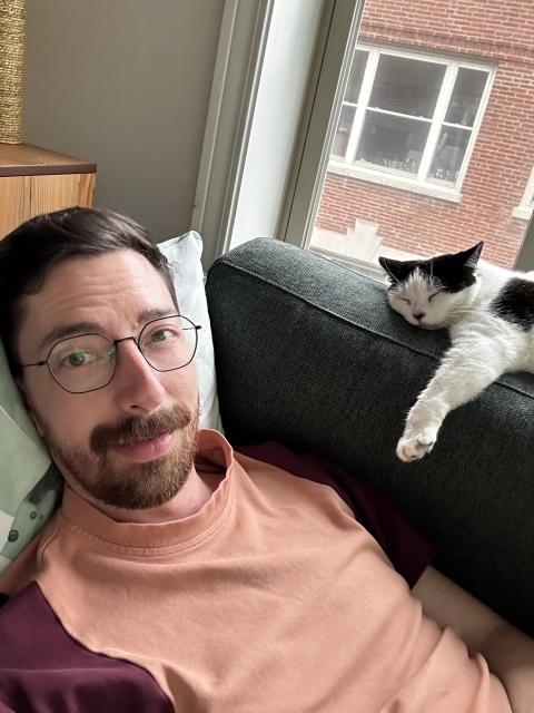 A bearded man lays on the couch, a black and white cat sleeps on the back cushion with a paw sticking out