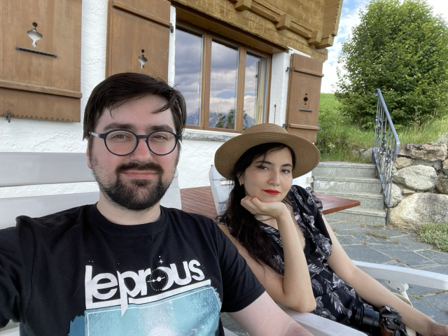 A selfie taken in front of a cabin. Reflection in the window reveals mountains. On the left, a man with black hair and round glasses wearing a Leprous t-shirt. On the right, a woman in a dark dress and a straw hat. 