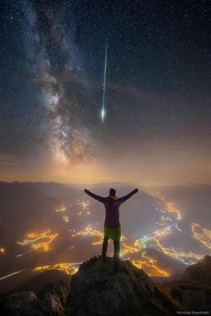 A person is seen facing away, standing on a peak. Other mountain peaks surround them. City lights are seen in towns and along roads below. Stars in the night sky are above. The band of the Milky Way galaxy slants down from the upper left. A bright green meteor streak slants down from above.