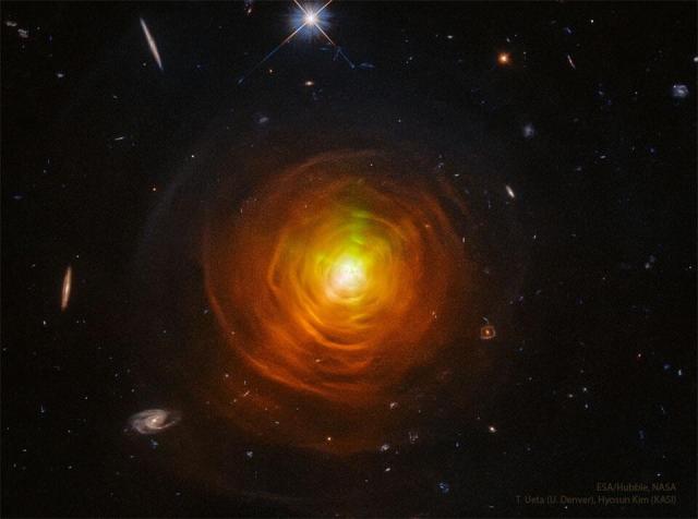 A star surrounded by orange shells and arcs sit in the center of a dark starfield. Galaxies from the distant universe can be seen around the edges.