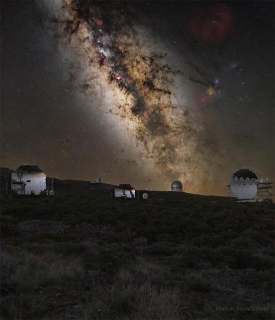 A mountaintop is shown covered by brush. Across the horizon are several telescopes. Behind the mountaintop is a deep exposure of the sky showing the central band of our Milky Way galaxy and several well-known stars and nebulas.