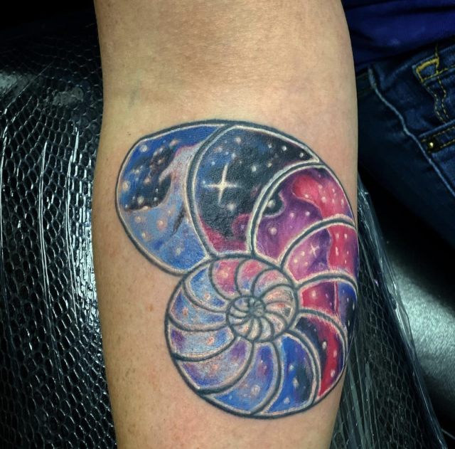 Forearm tattoo of an ammonite shell with pink, blue, and purple clouds, and white stars - similar to a nebula.