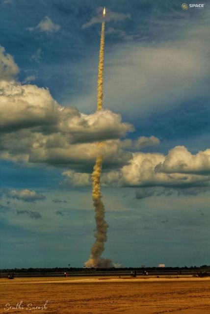 A rocket is seen after lift-off with a long smoke plume. The rocket is captured against a blue sky and has gone through a cloud deck. In the foreground is an empty tan-colored field.