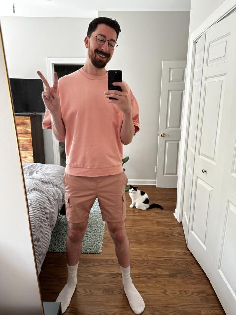 A tall bearded man stands in front of a mirror wearing a pink short sleeve sweatshirt and rose colored shorts while smiling and making a peace sign. A cat sits in the background of the room.