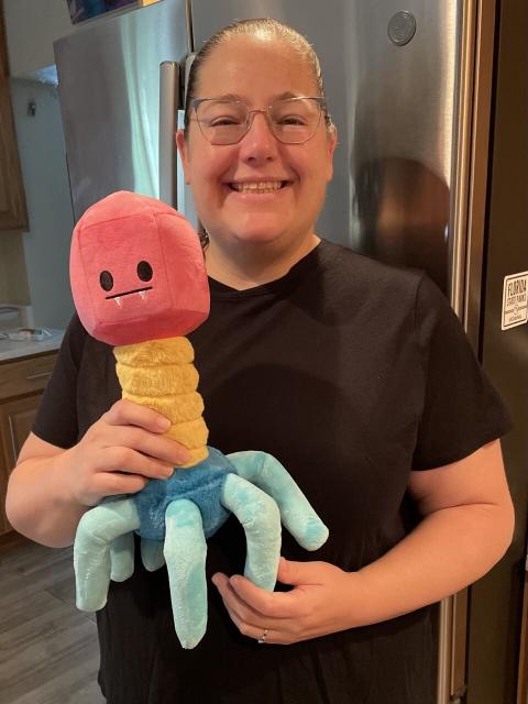 Photo of a woman (me) holding a giant plush phage (virus that infects bacteria). The phage has a red head (with adorable fangs), yellow neck and blue tail 