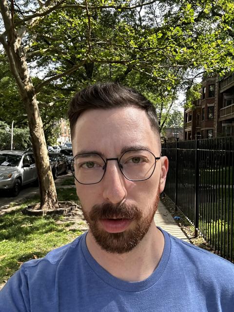 A man with a beard and glasses stands outside and looks into the camera showing off his haircut. He wears a blue t-shirt and there is green grass and a lush tree in the background. 