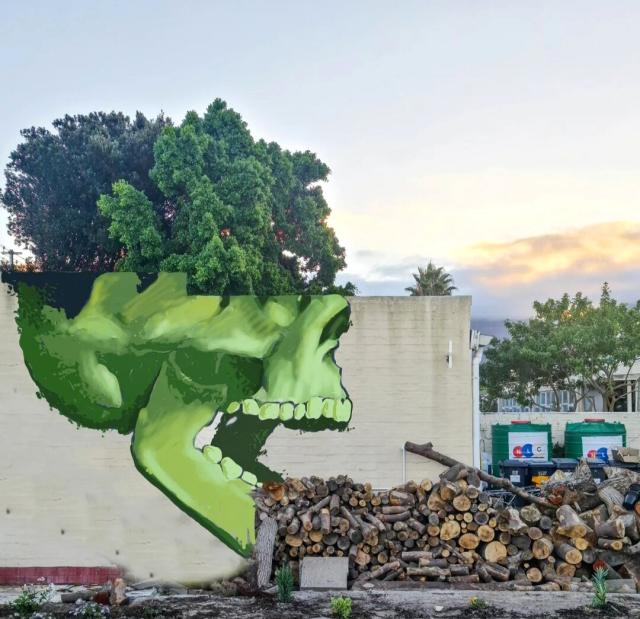 Streetart. A green skeleton head is painted / sprayed on a beige street wall.The head is depicted from the side and has an open mouth. In front of the wall is a long stack of logs lined up. Behind the wall is a tree with green leaves. The whole thing looks like a green skeleton skull with green hair eating a row of logs.
Title: "You are what you eat"