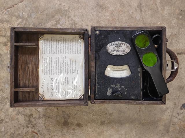 A strange device in a dark wooden case. It has a wand with two green discs visible. There is a metal plaque which reads, "MacAffy's Anti-Viral 'Revigorator'" and a vintage-seeming pamphlet with usage instructions. The device has a readout for "foot-candles."