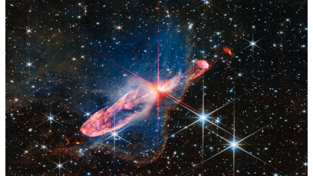 From JWST: At the center is a thin horizontal orange cloud known as Herbig-Haro 46/47 that is uneven with rounded ends, and tilted from bottom left to top right. It takes up about two-thirds of the length of this angle, but is thin at the opposite angle. At its center is a red-and-pink star with prominent, eight-pointed diffraction spikes. It has a central yellow-white blob. The orange lobe to the left is fatter. Just off the edge is a tiny red arc that curves in the opposite direction. The right lobe is thinner, and ends in a smaller orange semi-circle that has a faint purple outline. Just off the edge of this lobe is a slightly smaller orange sponge-like blob. A thin, undulating blue line runs from the central stars through the right lobe. A delicate, semi-transparent blue cloud known as a nebula drifts toward the top of the image and peters out toward the left of the frame, but toward the right and bottom, it ends in a soft ridge set off in a translucent orange. The background is filled with stars and galaxies.
