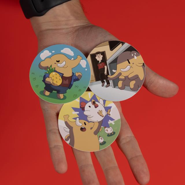 Three stickers on top of a person's palm. One sticker features an elephant dressed in a jean outfit and holding a pineapple. Another features an elephant hiding under a table while a man in a red space outfit is looking for him. The last one features an elephant holding a chicken, with more chickens in the background.