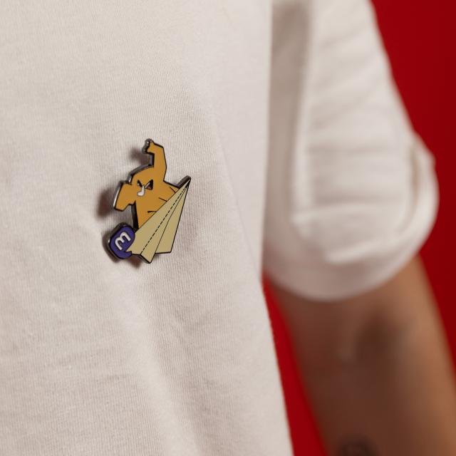An enamel pin attached to a white t-shirt. The pin features an elephant riding a paper airplane.