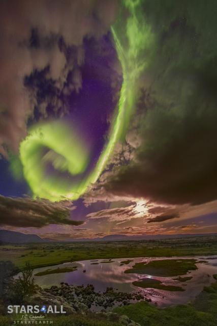 A bright green spiral aurora is seen in a break in the clouds before a purple background. The foreground contains green grassland and a circular lake.