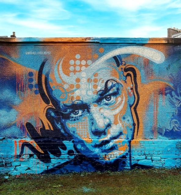 Streetartwall. On a long wall is sprayed/painted a mural of a man in pop art graffiti style. The background is blue tones with white and orange fog waves. On it appears a bald young man whose face is decorated with orange and white dots, grids and waves. The look of the beautiful blue man goes directly to us.