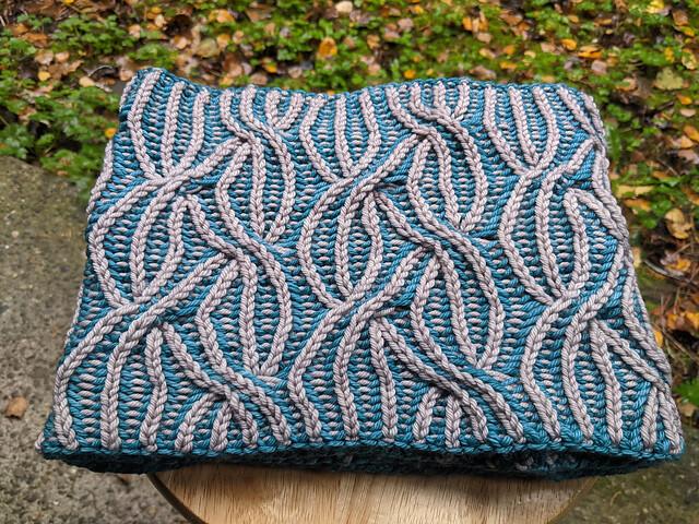 The "inside" of a two color teal and grey cabled brioche cowl. It appears as a simple square with overlapping "stripes" of grey vertical on a teal background.