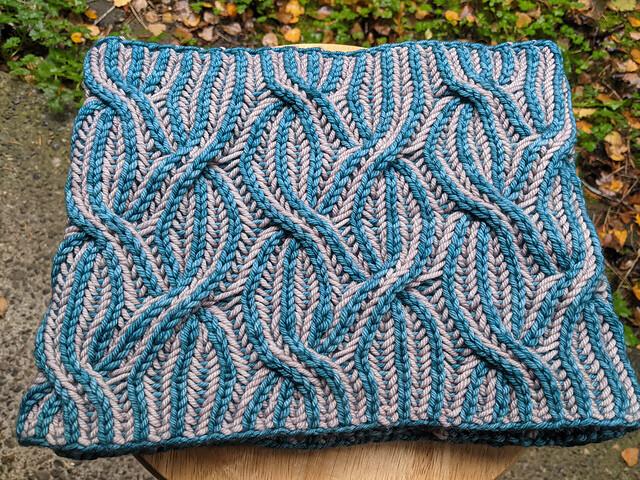 The "outside" of a cabled teal and grey brioche cowl. It appears as a simple square with overlapping "stripes" of teal vertical on a grey background.