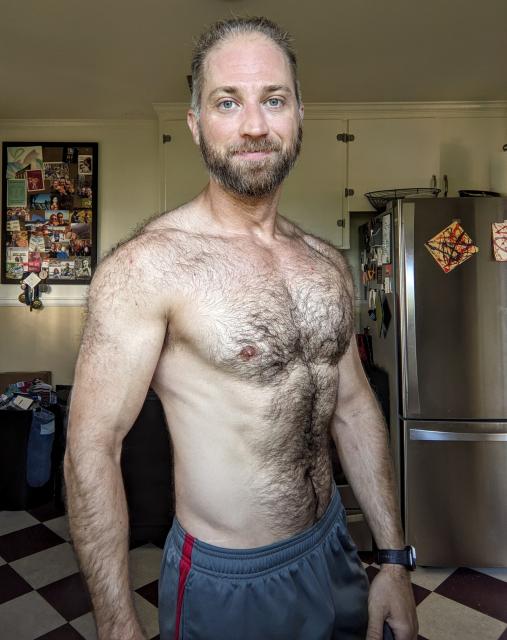 Shirtless furry-chested man stands in a kitchen ... smirking? smiling? 
