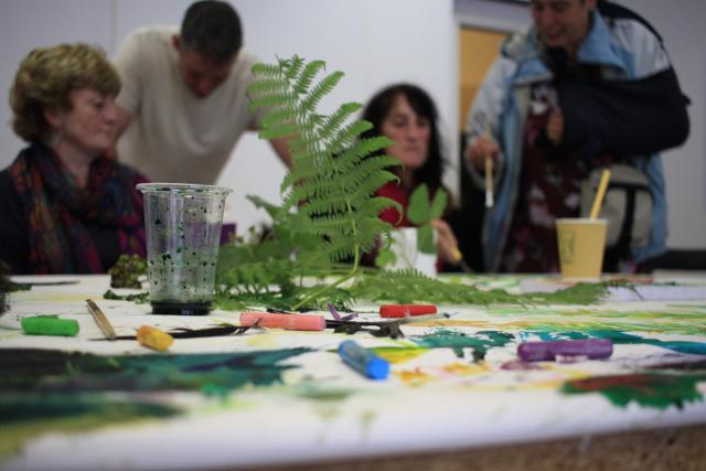 People work on a collaborative painting in the background, while the foreground shows ferns, branches and pastels on a paper sheet across a table