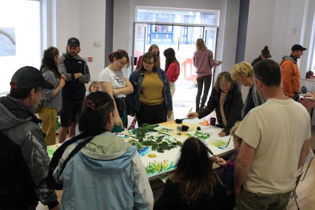 A large crowd attend a social event, sharing space and making art collectively. In the background an open door leads out onto a street.