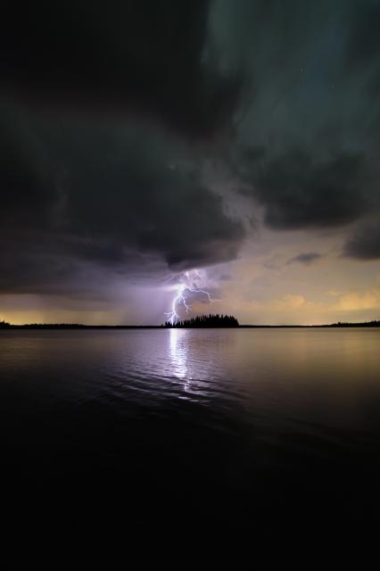A blue-white lightning strike extends down from low-hanging, green-grey clouds at night, appearing to strike just behind a small forested island in centre distance. The clouds break across a green-fading-to-gold sky, with a rippling lake below reflecting back the sky and the bolt of lightning.