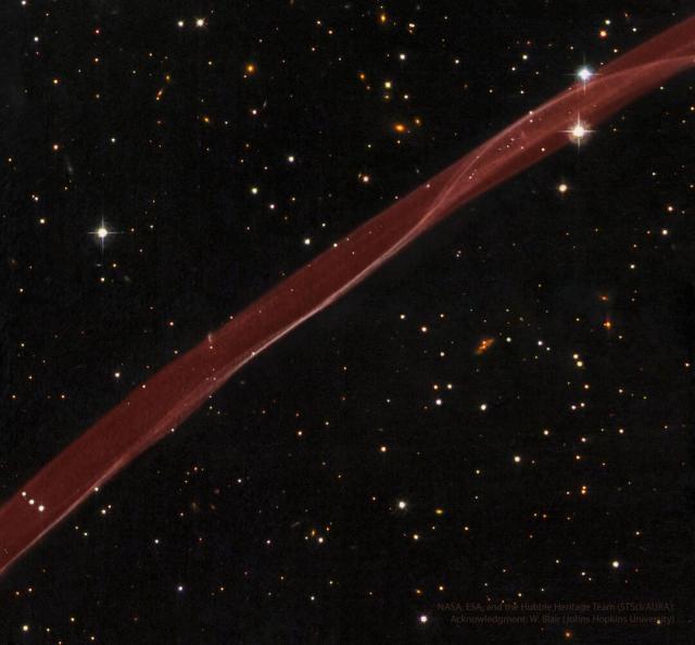 A thick transparent ribbon of red gas runs from the lower left to the upper right. A dark starfield with stars and galaxies surrounds the bright red ribbon.