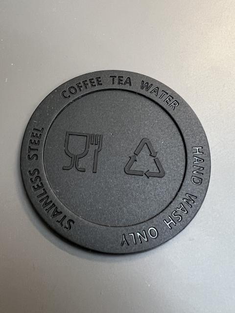 A flat circular piece of plastic with a raised edge. Around the edge is the text “Stainless steel” “coffee tea water” and “hand wash only”, equally spaced. In the centre recess are a symbol of a glass and fork, and a triangle made up of bent arrows.
