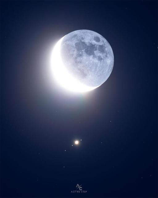 Earth Moon, in crescent phase, is seen just above the image center. Directly below is a bright spot surrounded by four other spots, all in a row, which are all moons of Jupiter.