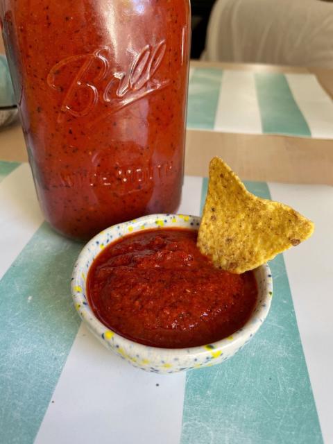 Small dish containing a portion of a dense dark red sauce, with a nacho inserted on it. Behind there’s the jar with all of the sauce
