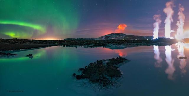 A body of water is seen in front of a night sky. The water reflects the sky. In the sky, on the right are green aurora. In the center is an orange plume. On the right are three while plumes.