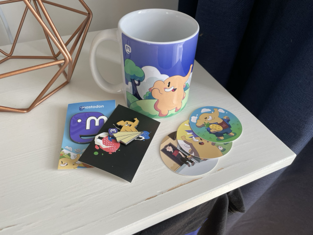 Mastodon mug, enamel pins, and stickers next to each other on a bedside table