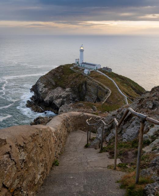 A flight of steps leads down a cliff to a white painted lighthouse. The sky is dark and the light from the lighthouse is visible against the sea backdrop.