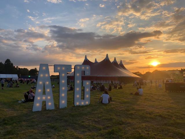 a photo of arctangent festival: in the front three large letters: ATG in white with light in them stand before a large red festival tent with the sunset behind it. 
there are people sitting in the grass and walking around. 