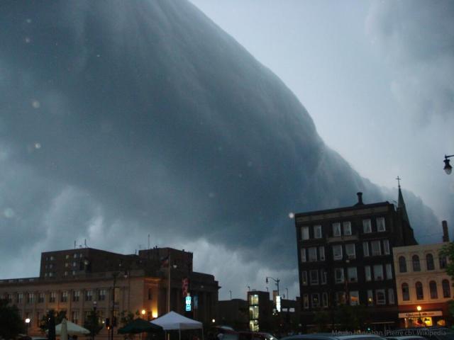 Building in a city are pictured. Above the buildings appears a long dark cylindrical cloud that goes to the horizon.