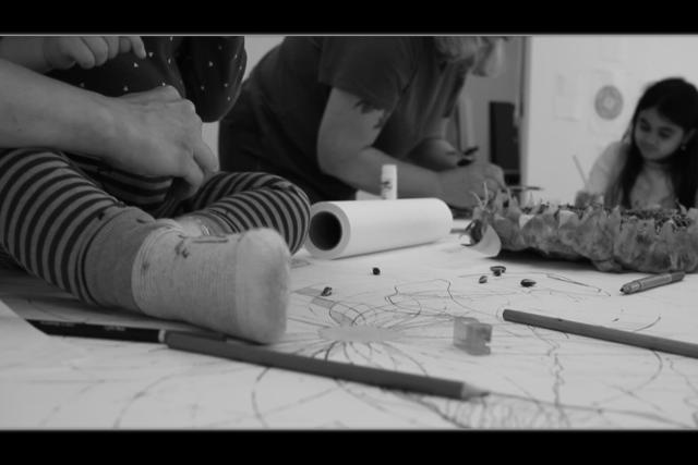 People are making art on a table with markers and paper, a roll of paper and a giant sunflower head visible. A baby's foot is in the foreground and an obscured child drawing in the background.