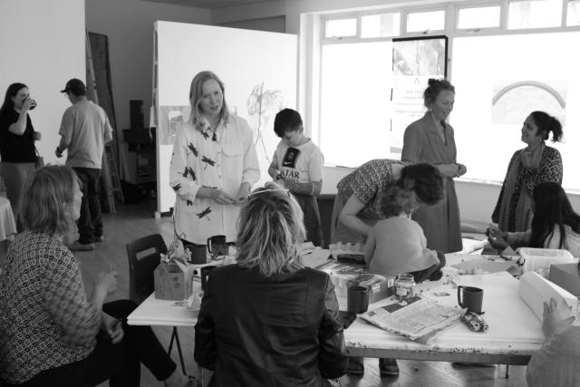 A large group of people are making art together and conversing over food and drinks in a brightly lit room.