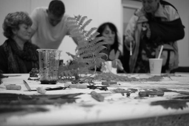 People sitting at a table conversing, obscured by fern leaves and art materials on a table. The people are out of focus.