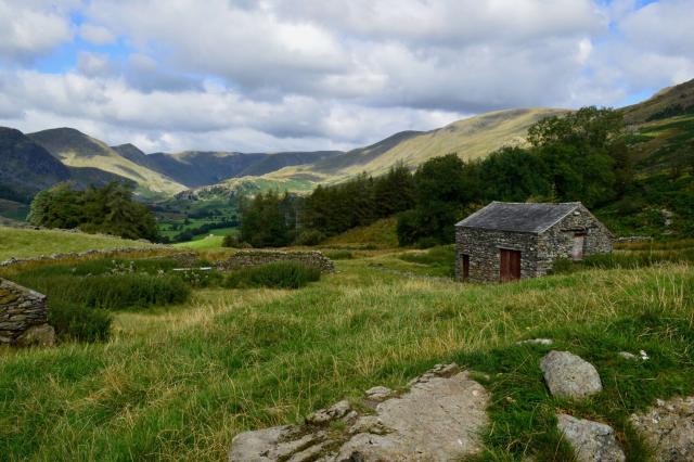 A ring of hills dappled by patchy sunlight, small woods and rocky outcrops. An old stone barn in the foreground with a red painted door.