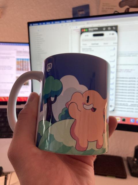 A hand holds a mug in front of a computer screen. The Mastodon-logo and an elephant/mastodon is printes on that very mug.