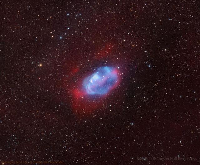 A diffuse nebula is seen against a dark starfield. The center of the nebula is blue and it is surrounded by a red glow.