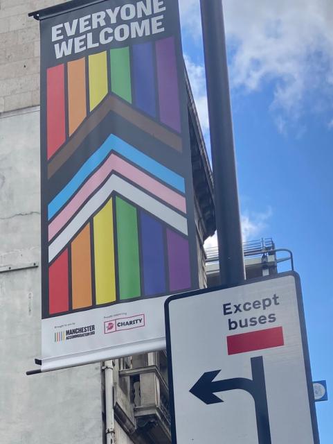 A banner on a street lamp saying “everyone welcome” with the Pride flag colours below, right next to a traffic sign that says “except buses”