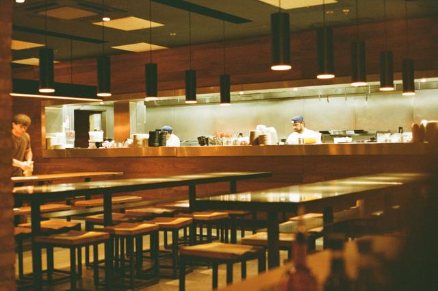 An empty restaurant hall illuminated by warm yellow light, with a sharp window to the kitchen, illuminated by bright white light
