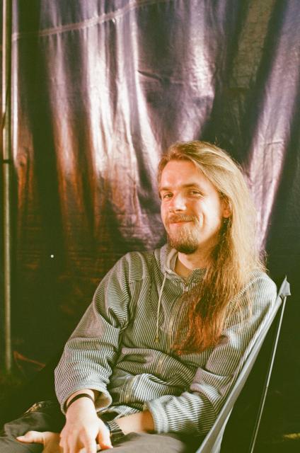A smiling man with long luscious blonde hair, mustache, and beard, illuminated partly by daylight, partly by stage lights