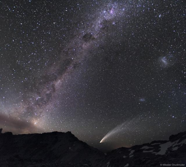 A rocky landscape is capped by a dark night sky. In the sky, the band of our Milky Way Galaxy runs along the right, while two fuzzy patches that are the LMC and SMC are visible on the right. Thousands of stars are resolved all over the frame.