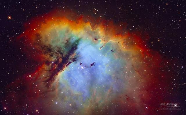 A nebula that appears blue in the middle and is surrounded by red-glowing gas is featured. Dramatic lanes of dark dust cut through the nebula's left side. A group of stars is visible toward the nebula's center.