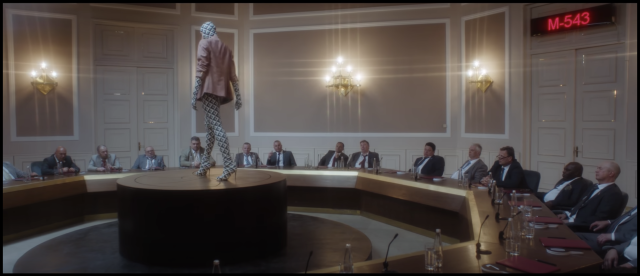 Still from the music video “Boom Boom.” 

In the scene a woman in a full body suit that covers her head, high heels, and a blazer with the sleeves pushed up, stands atop a dais in the center of a many-sided table. 

Around the table are seated many men wearing suits, forming a panel seemingly convened to judge women.