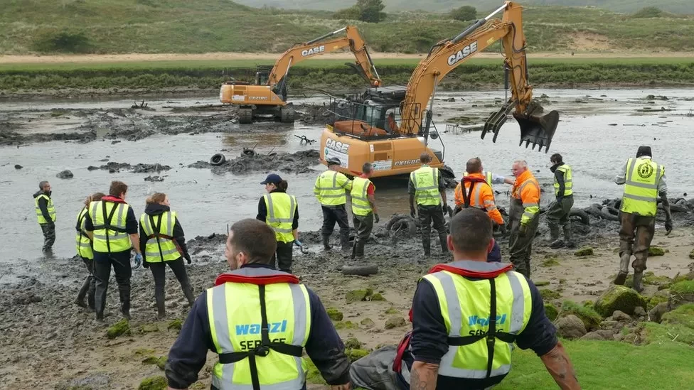 Several people in high vis and two diggers dredging a muddy river.