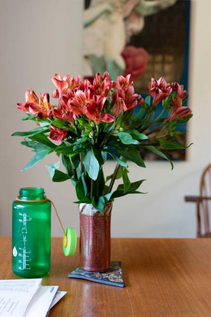On a table: flowers in an earthenware vase, a Nalgene bottle, and, a stack of incoming mail.