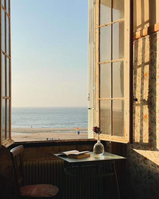 A colour photo of casement windows, open over a table that has on it a book and a single red flower in a clear, bulbous glass vase. The window looks out onto the sea and a wide beach with a few distant human figures. 