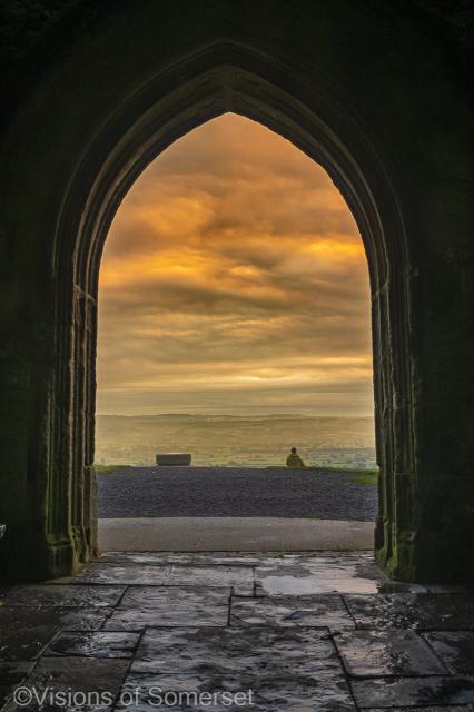 A lone figure sits watching sunrise. The view is through the archway. Golden clouds above the land.