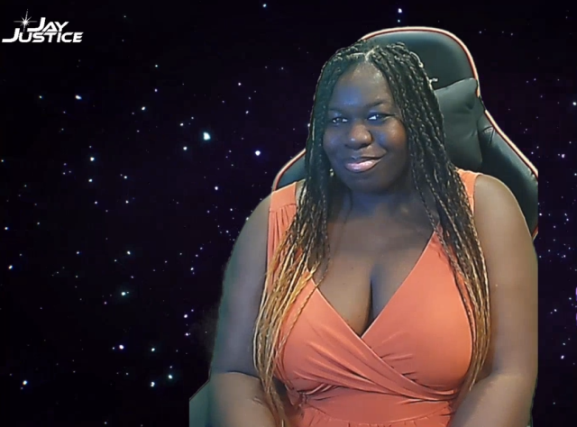 a smiling Black person with long blonde and black braids wearing an orange dress, sitting in front of a galaxy background