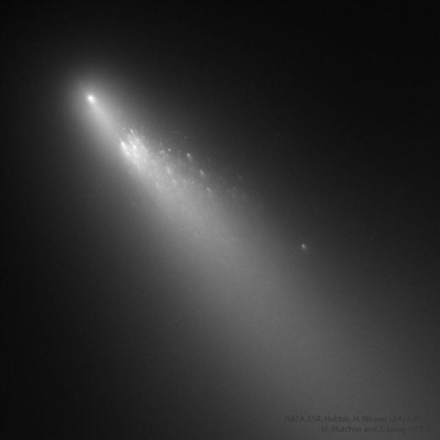 A fuzzy comet is shown in gray on the upper left against a dark space background. The comet's tail extends diagnonally to the lower right. The main part of the comet is seen broken up into many trailing pieces.